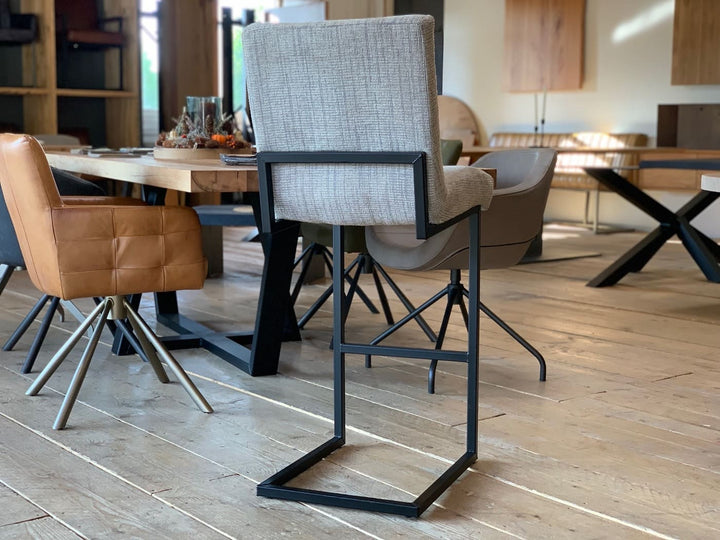 Barhocker Stoffbezug Industrial Rückansicht: Moderner Barhocker mit hellgrauem Stoffbezug, gepolsterter Rückenlehne und Sitzfläche sowie schwarzem Metallgestell im Industrial-Stil, platziert auf einem Holzboden in einem stilvollen Möbelgeschäft mit weiteren Sitzmöbeln im Hintergrund.