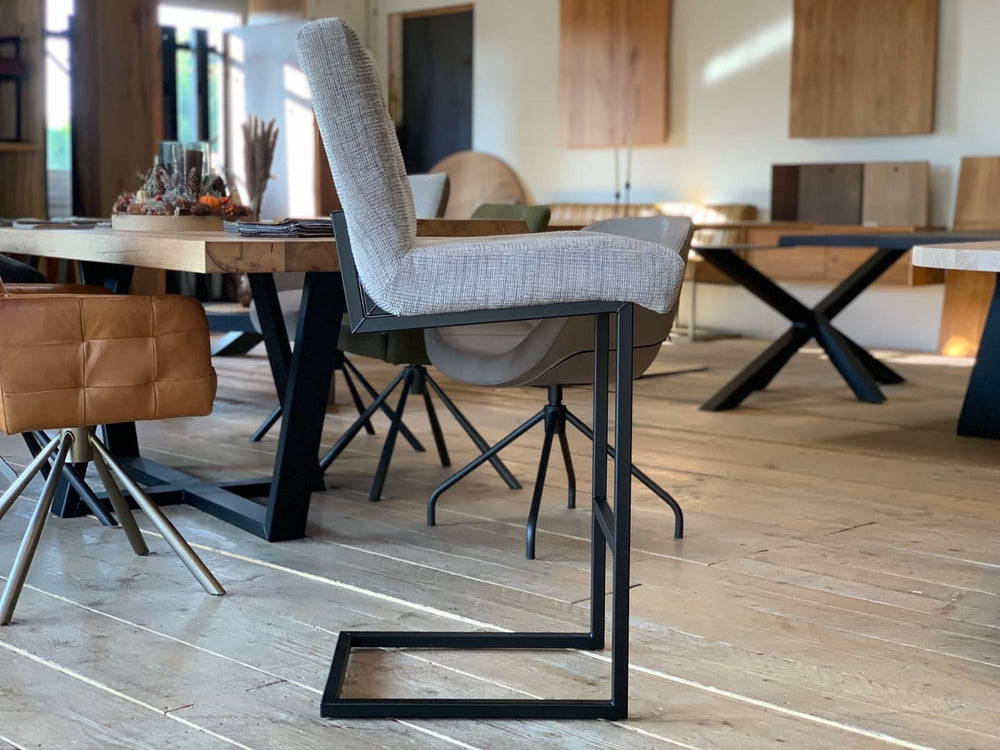Barhocker Stoffbezug Industrial Seitenansicht: Moderner Barhocker mit hellgrauem Stoffbezug, gepolsterter Sitzfläche und Rückenlehne sowie schwarzem Metallgestell im Industrial-Stil, platziert auf einem Holzboden in einem stilvollen Möbelgeschäft mit weiteren Sitzmöbeln im Hintergrund.