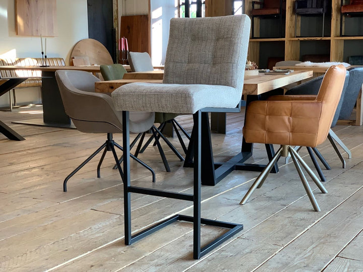 Barhocker Stoffbezug Industrial: Moderner Barhocker mit hellgrauem Stoffbezug, gepolsterter Sitzfläche und Rückenlehne, kombiniert mit einem schwarzen Metallgestell im Industrial-Stil, präsentiert auf einem Holzboden in einem stilvollen Möbelgeschäft mit weiteren Sitzmöbeln im Hintergrund.