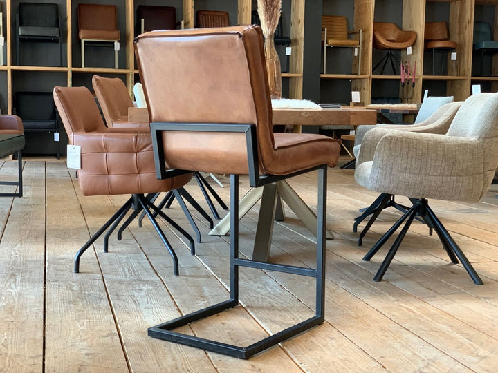 Barstuhl Leder Industrial in Rückansicht: Brauner Lederstuhl mit gepolsterter Rückenlehne und Sitzfläche sowie schwarzem Metallgestell im Industrial-Stil, platziert auf einem Holzboden in einem stilvollen Möbelgeschäft mit weiteren Stühlen und einem Tisch im Hintergrund.