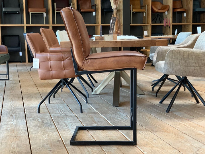 Barstuhl Leder Industrial in Seitenansicht: Brauner Lederstuhl mit gepolsterter Sitzfläche und Rückenlehne, getragen von einem schwarzen Metallgestell im Industrial-Stil, steht auf einem Holzboden in einem modernen Möbelgeschäft mit weiteren Stühlen und einem Tisch im Hintergrund.