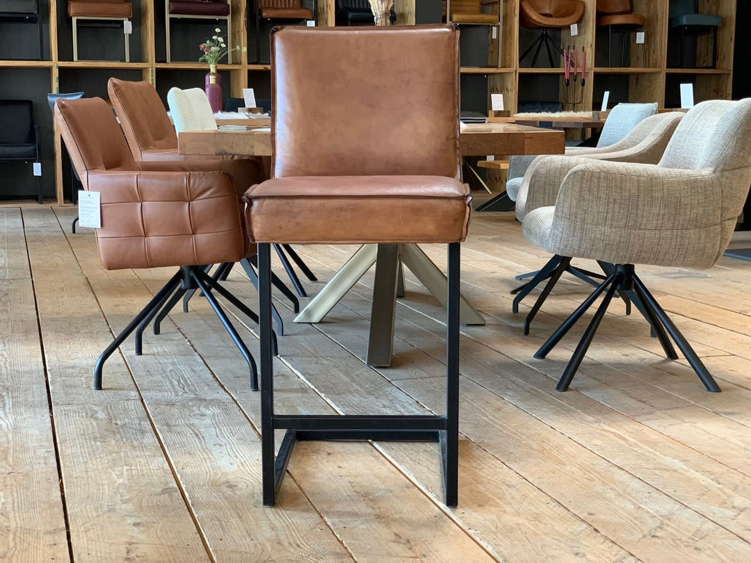 Barstuhl Leder Industrial in Vorderansicht: Brauner Leder-Barstuhl mit gepolsterter Sitzfläche und hoher Rückenlehne auf einem schwarzen Metallgestell im Industrial-Stil, präsentiert auf einem Holzboden in einem modernen Möbelgeschäft mit weiteren Sitzmöbeln im Hintergrund.