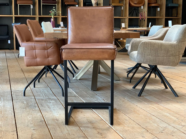 Barstuhl Leder Industrial in Vorderansicht: Brauner Leder-Barstuhl mit gepolsterter Sitzfläche und hoher Rückenlehne auf einem schwarzen Metallgestell im Industrial-Stil, präsentiert auf einem Holzboden in einem modernen Möbelgeschäft mit weiteren Sitzmöbeln im Hintergrund.
