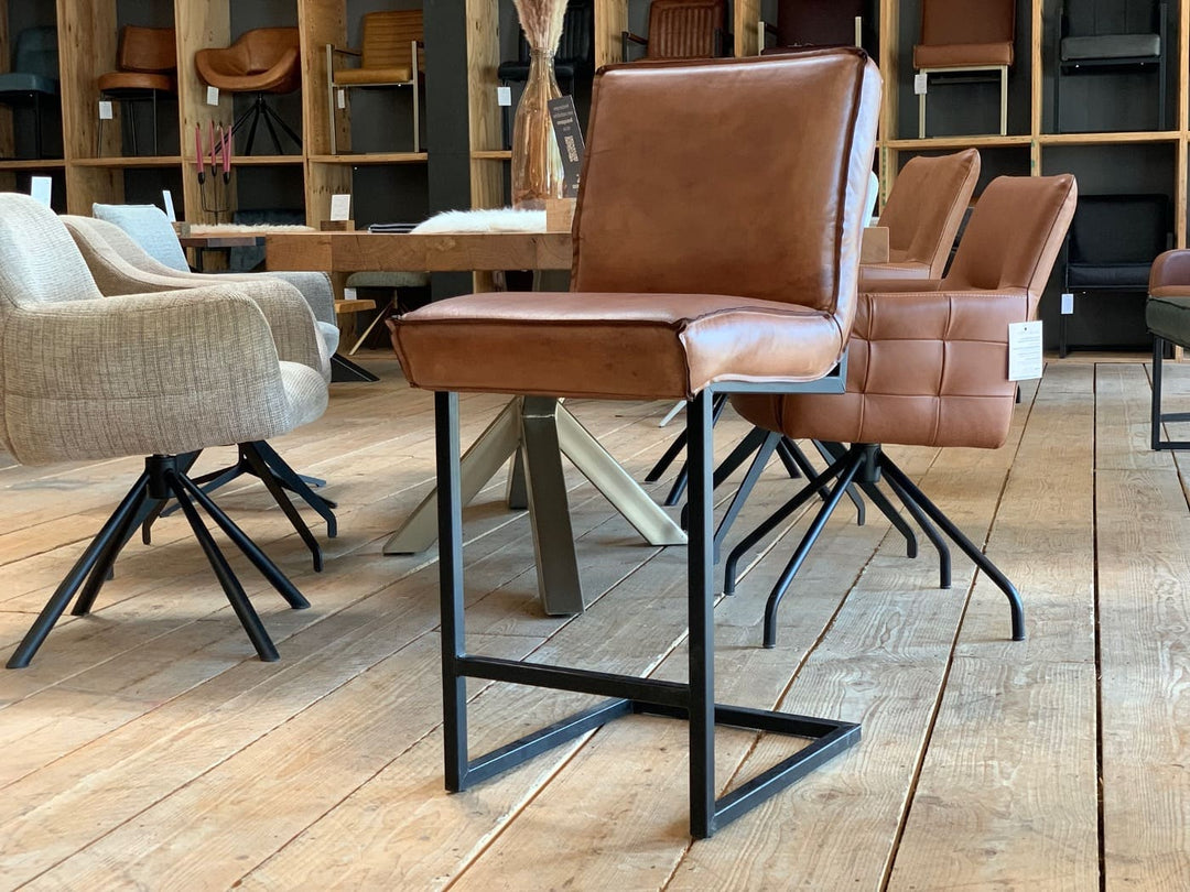 Barstuhl Leder Industrial im modernen Möbelgeschäft: Ein hochwertiger, brauner Leder-Barstuhl mit klaren Linien und schwarzem Metallgestell im Industrial-Stil steht auf einem Holzfußboden, umgeben von weiteren Stühlen und einem Esstisch im Hintergrund.