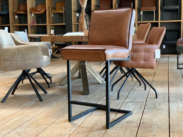 Barstuhl Leder Industrial im modernen Möbelgeschäft: Ein hochwertiger, brauner Leder-Barstuhl mit klaren Linien und schwarzem Metallgestell im Industrial-Stil steht auf einem Holzfußboden, umgeben von weiteren Stühlen und einem Esstisch im Hintergrund.
