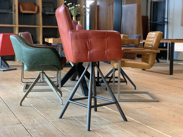Barstuhl drehbar Leder Industrial Seitenansicht: Moderner, drehbarer Barstuhl aus rotem Leder mit gepolsterter Sitzfläche, Rückenlehne und Armlehnen, kombiniert mit einem stabilen Metallgestell im Industrial-Stil, präsentiert in einem stilvollen Möbelgeschäft mit weiteren Sitzmöbeln im Hintergrund.