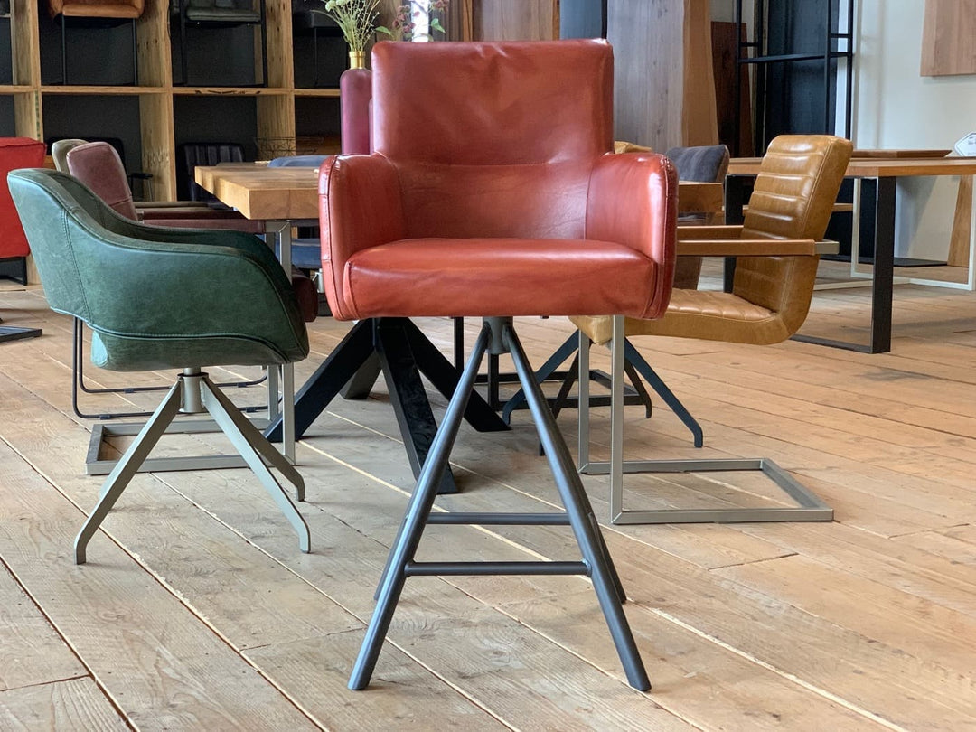 Barstuhl drehbar Leder Industrial Vorderansicht: Moderner, drehbarer Barstuhl aus rotem Leder mit gepolsterter Sitzfläche, Rückenlehne und Armlehnen, kombiniert mit einem stabilen Metallgestell im Industrial-Stil, präsentiert in einem stilvollen Möbelgeschäft mit weiteren Sitzmöbeln im Hintergrund.