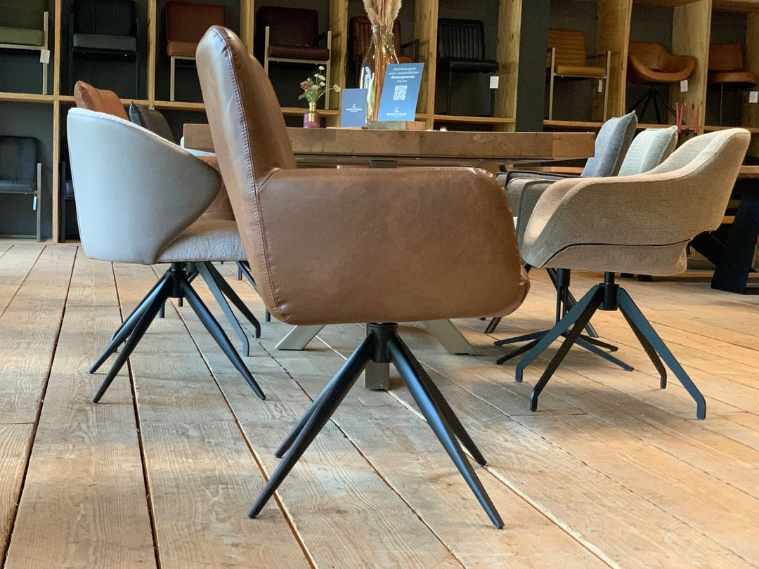 Seitliche Ansicht eines modernen Büffelleder Drehstuhl in brauner Ausführung mit gepolsterter Sitzfläche, hoher Rückenlehne und Armlehnen auf schwarzem Metall-Drehgestell, platziert an einem Holztisch mit weiteren stilvollen Stühlen in einem wohnlichen Ambiente.