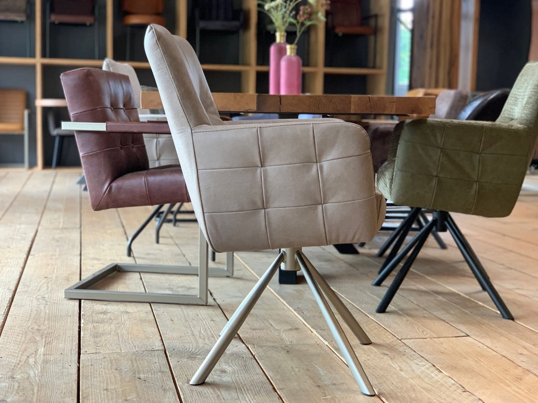 Seitliche Ansicht eines modernen Drehstuhl Leder hohe Rückenlehne in hochwertigem Leder in der Farbe Taupe, mit gepolsterter Sitzfläche, Armlehnen und dekorativer Steppung auf einem stabilen Metall-Drehgestell, platziert an einem Holztisch in einem stilvollen Essbereich.