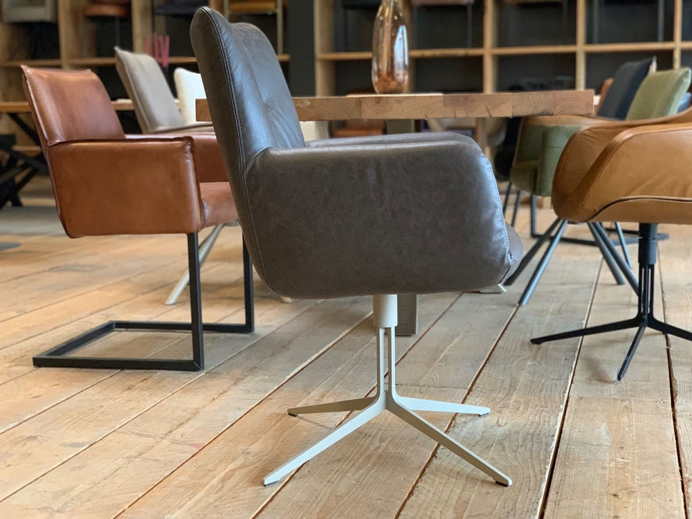 Seitliche Ansicht eines modernen Esszimmerstuhl Drehstuhl in grauer Lederoptik mit gepolsterter Sitzfläche, hoher Rückenlehne und Armlehnen auf einem hellen Metall-Drehgestell, platziert an einem Holztisch mit weiteren stilvollen Stühlen in einem wohnlichen Ambiente.