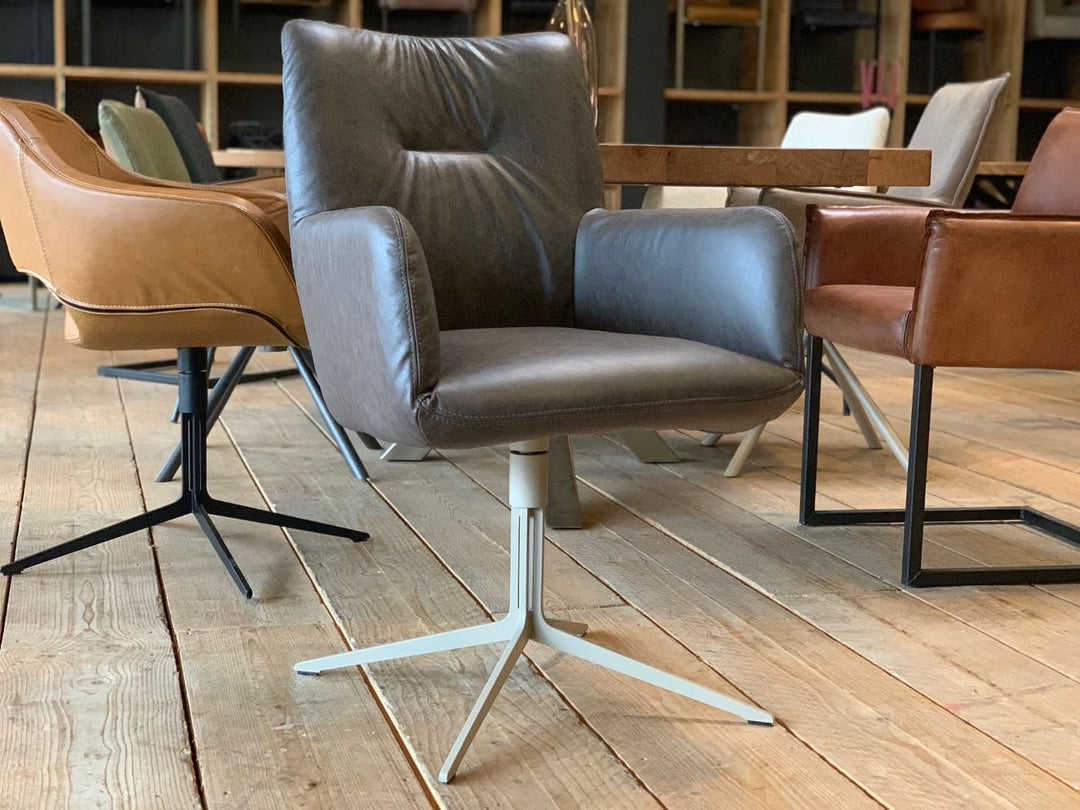 Moderner Esszimmerstuhl Drehstuhl in grauer Lederoptik mit gepolsterter Sitzfläche, hoher Rückenlehne und Armlehnen auf einem hellen Metall-Drehgestell, platziert an einem Holztisch und kombiniert mit weiteren stilvollen Stühlen in einem wohnlichen Ambiente.