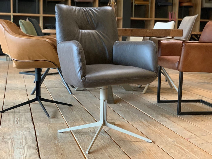 Moderner Esszimmerstuhl Drehstuhl in grauer Lederoptik mit gepolsterter Sitzfläche, hoher Rückenlehne und Armlehnen auf einem hellen Metall-Drehgestell, platziert an einem Holztisch und kombiniert mit weiteren stilvollen Stühlen in einem wohnlichen Ambiente.