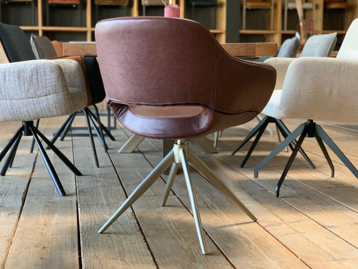 Rückansicht eines eleganten Esszimmerstuhl Leder drehbar-182T mit brauner Lederoberfläche, weich gepolsterter Rückenlehne und stilvoller Nahtführung, ausgestattet mit einem drehbaren Metallgestell in moderner Sternform; positioniert an einem Holztisch und umgeben von weiteren Esszimmerstühlen in einem wohnlichen Ambiente.