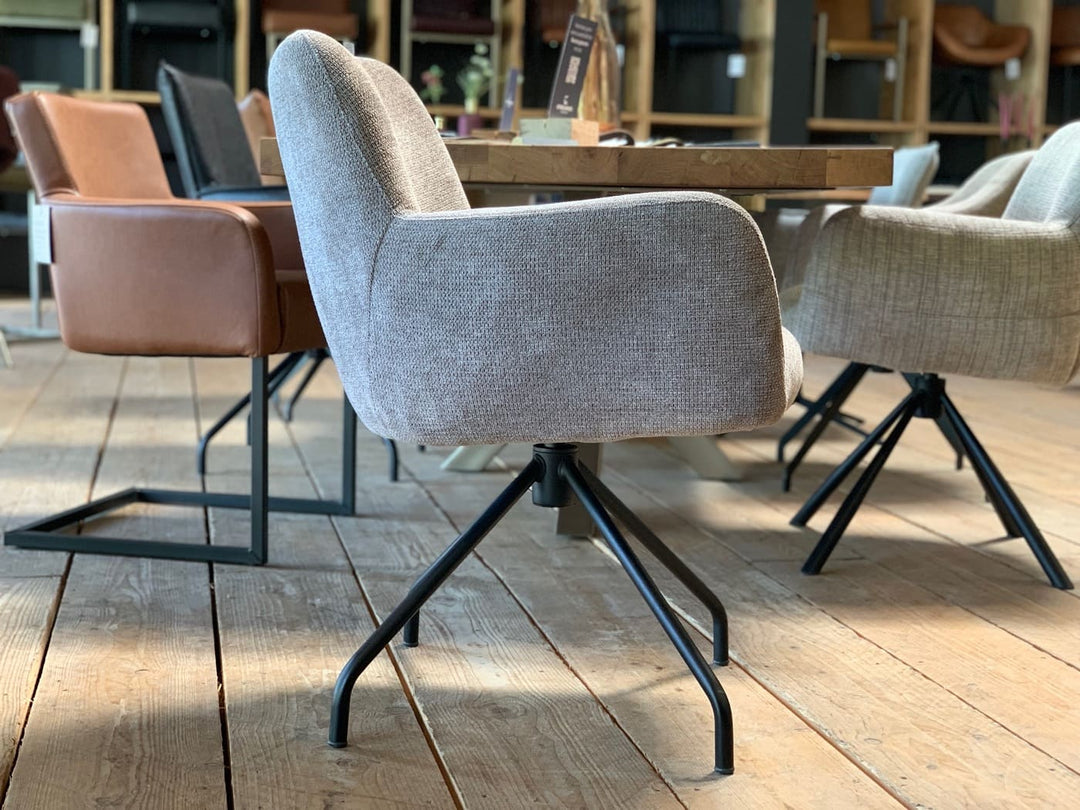Gepolsterter Drehstuhl Esszimmer in Seitenansicht mit grauem Stoffbezug, ergonomischer Sitzschale und Armlehnen, drehbar auf schwarzem Stahlgestell mit stabilen, schräg stehenden Beinen, in modernem Essbereich mit Holztisch.