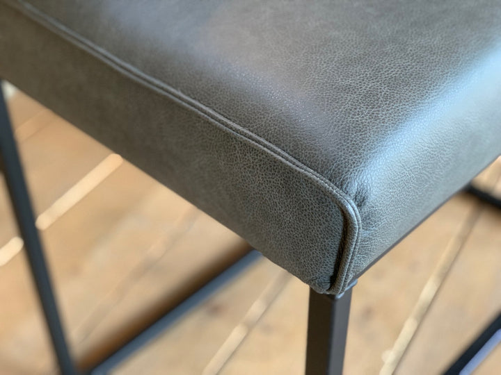 Detailaufnahme einer Hochbank aus Leder mit grauem Sitzpolster, feiner Naht und sichtbarer Lederstruktur, kombiniert mit schwarzem Metallgestell im modernen Design.