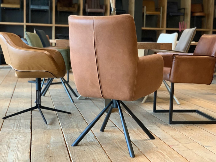 Rückansicht eines Leder Esszimmer Drehstuhl in Cognac mit hoher, leicht geschwungener Rückenlehne und gepolsterter Sitzfläche auf schwarzem Metall-Drehgestell, platziert an einem Holztisch mit weiteren stilvollen Stühlen in einem wohnlichen Ambiente.