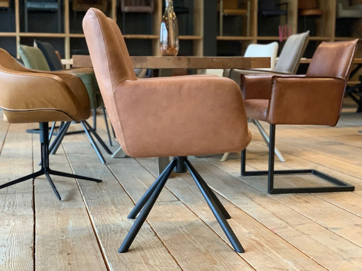 Seitliche Ansicht eines Leder Esszimmer Drehstuhl in Cognac mit gepolsterter Sitzfläche, hoher Rückenlehne und Armlehnen auf schwarzem Metall-Drehgestell, platziert an einem Holztisch mit weiteren stilvollen Stühlen in einem wohnlichen Ambiente.