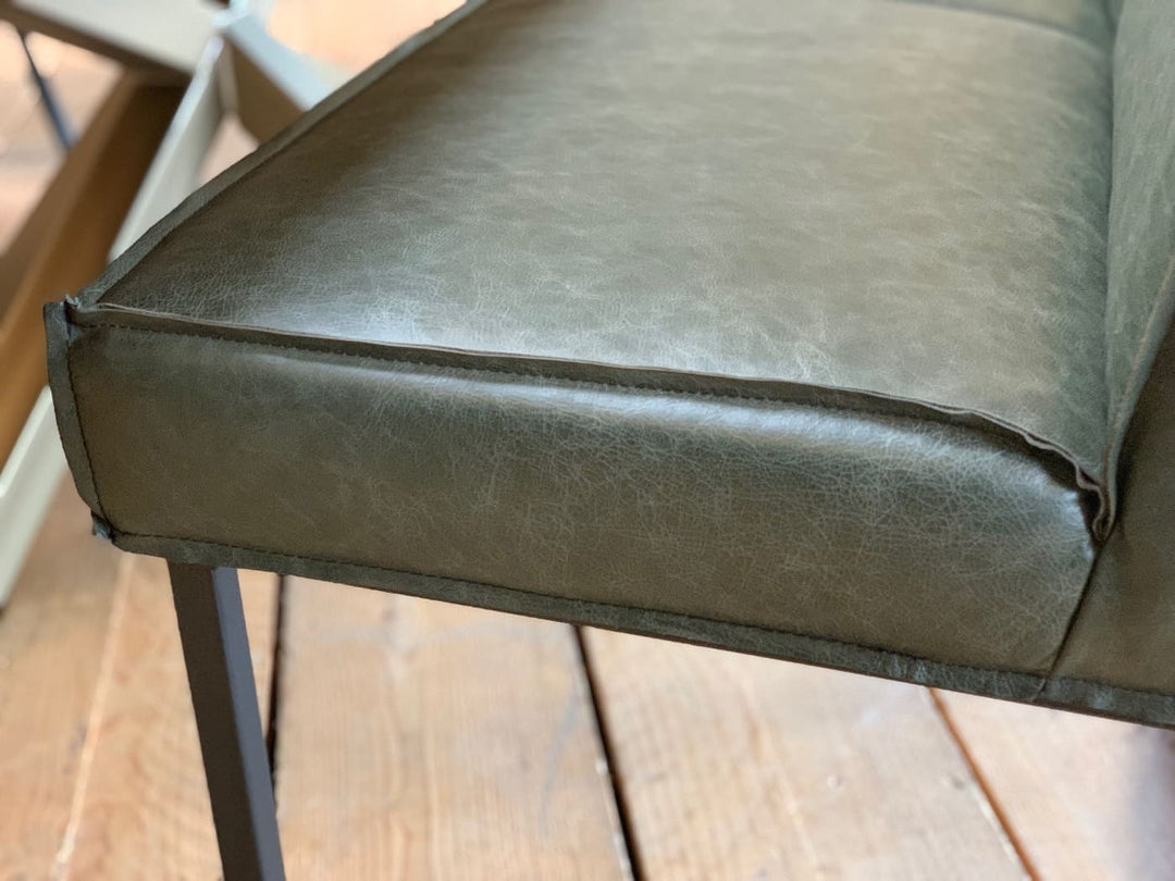 Detailaufnahme einer grünen Leder-Sitzbank mit sichtbarer Kedernaht am Sitzpolster, getragen von einem schwarzen Metallgestell auf Holzboden.