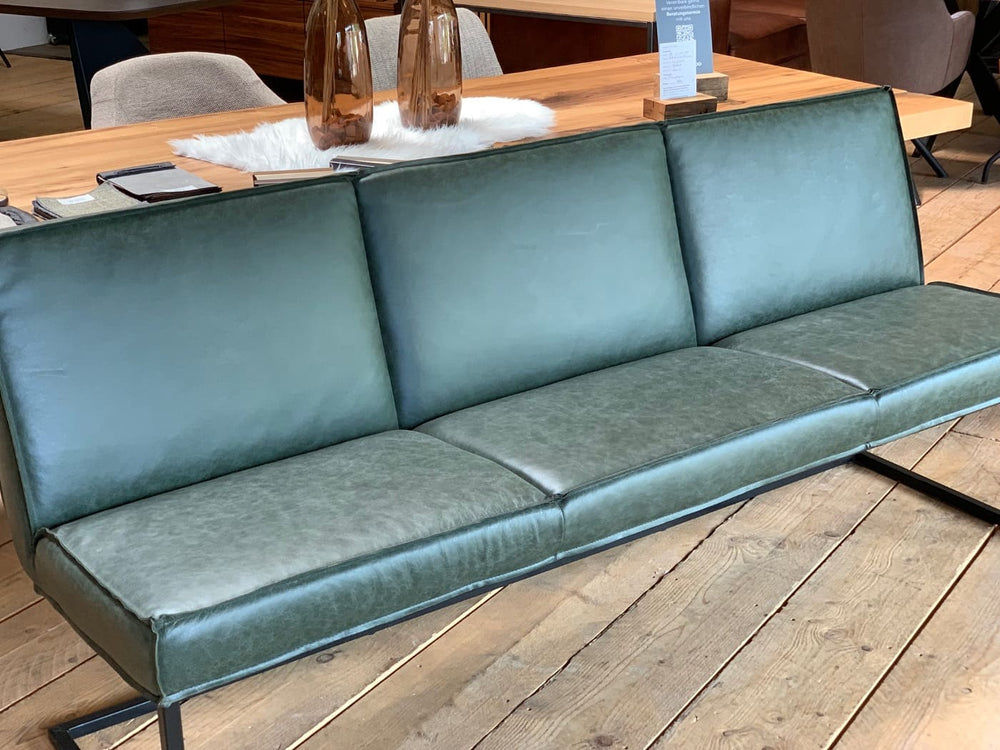 Schrägansicht einer modernen Sitzbank mit grünem Lederbezug und schwarzem Metallgestell vor einem Holztisch im Möbel-Showroom