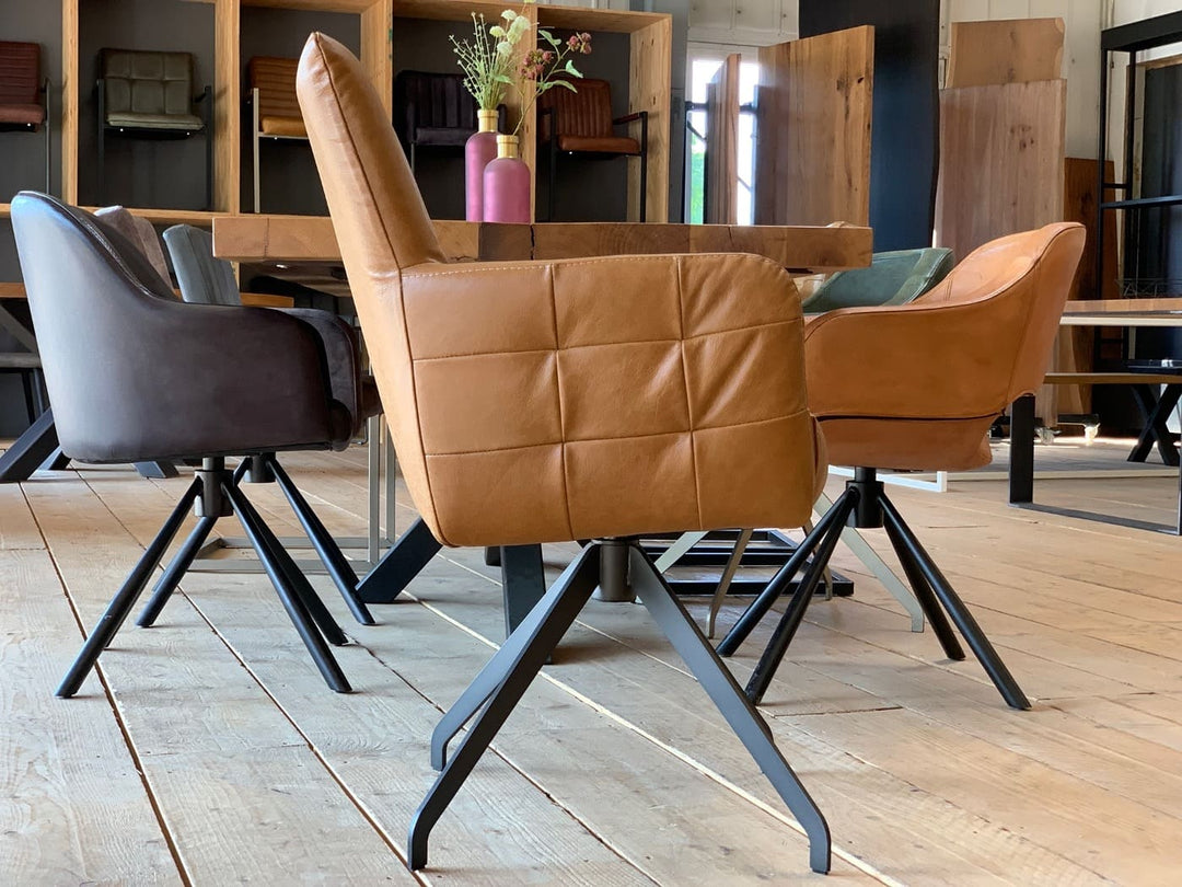 Seitliche Ansicht eines modernen Lederstuhl hohe Rückenlehne drehbar in der Lederfarbe Cognac mit gepolsterter Sitzfläche, Armlehnen und dekorativer Steppung auf schwarzem Metall-Drehgestell, platziert an einem Holztisch in einem stilvollen Essbereich.