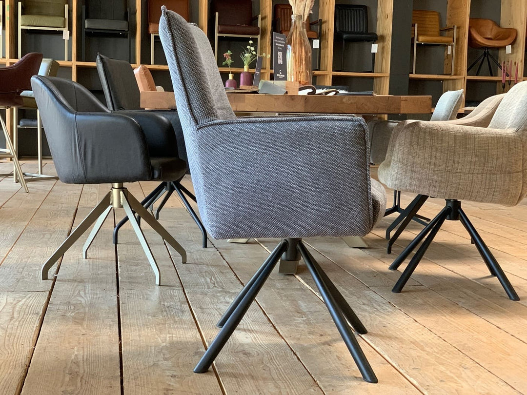 Moderner, drehbarer Esszimmerstuhl in grauem Stoff mit Armlehnen und hoher Rückenlehne, auf einem schwarzen Metall-Drehgestell. Seitenansicht in einem stilvollen Essbereich mit Holzboden, Tisch und weiteren gepolsterten Stühlen im Hintergrund.