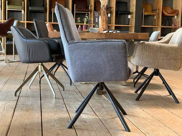 Moderner, drehbarer Esszimmerstuhl in grauem Stoff mit Armlehnen und hoher Rückenlehne, auf einem schwarzen Metall-Drehgestell. Seitenansicht in einem stilvollen Essbereich mit Holzboden, Tisch und weiteren gepolsterten Stühlen im Hintergrund.