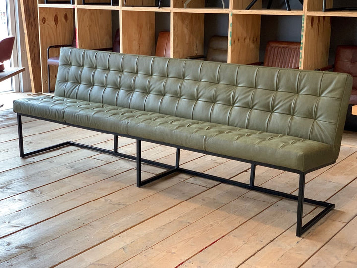 Moderne Polsterbank aus Leder für das Esszimmer mit gesteppter Sitz- und Rückenfläche in Olivgrün, auf schwarzem Metallgestell, stilvoll vor Holzboden und Regalwand präsentiert