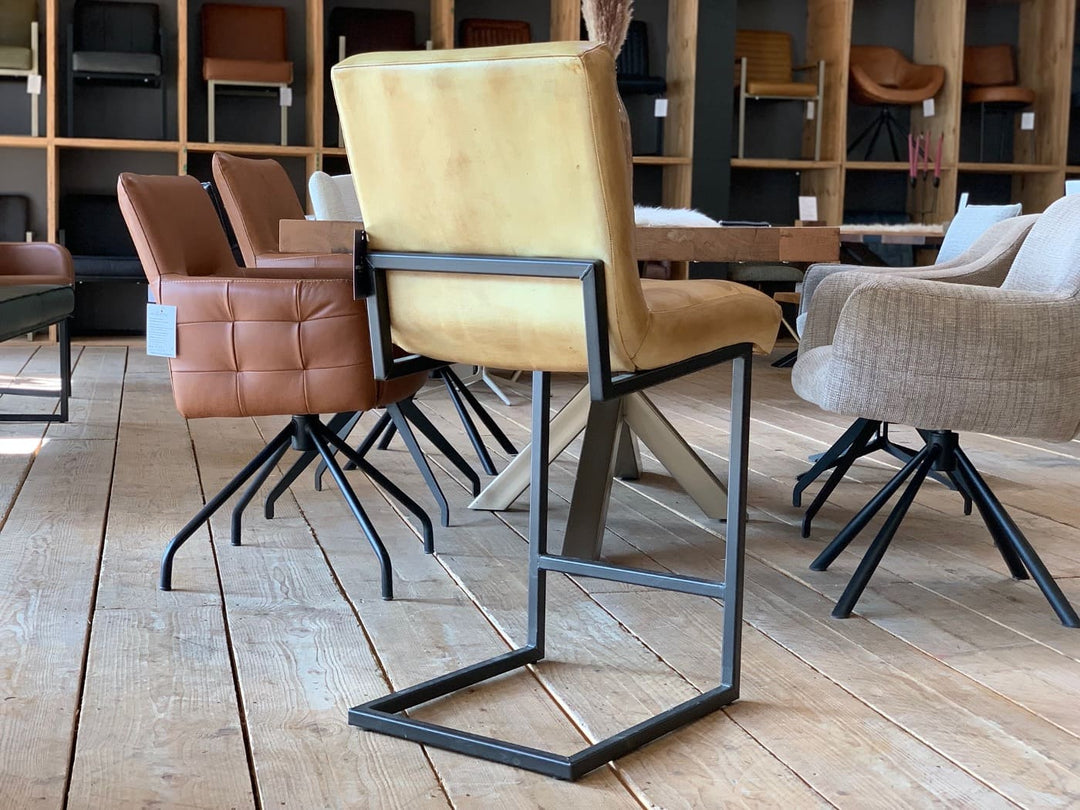 Thekenstuhl Leder Industrial in Mustard Rückansicht: Eleganter Thekenstuhl aus senfgelbem Leder mit gepolsterter Rückenlehne und Sitzfläche, kombiniert mit einem schwarzen Metallgestell im Industrial-Stil, platziert auf einem Holzboden in einem modernen Möbelgeschäft mit weiteren Sitzmöbeln im Hintergrund.