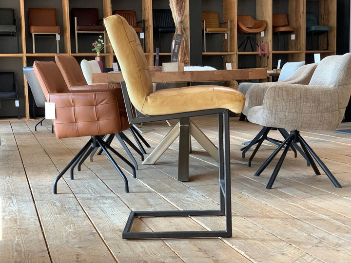 Thekenstuhl Leder Industrial in Mustard Seitenansicht: Moderner Thekenstuhl aus senfgelbem Leder mit gepolsterter Sitzfläche und Rückenlehne, getragen von einem schwarzen Metallgestell im Industrial-Stil, auf einem Holzboden in einem stilvollen Möbelgeschäft mit weiteren Sitzmöbeln im Hintergrund.