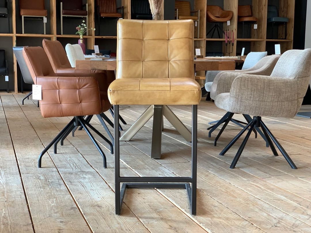 Thekenstuhl Leder Industrial in Mustard Vorderansicht: Moderner Thekenstuhl aus senfgelbem Leder mit gesteppter Rückenlehne und gepolsterter Sitzfläche, kombiniert mit einem schwarzen Metallgestell im Industrial-Stil, präsentiert auf einem Holzboden in einem stilvollen Möbelgeschäft mit weiteren Stühlen im Hintergrund.
