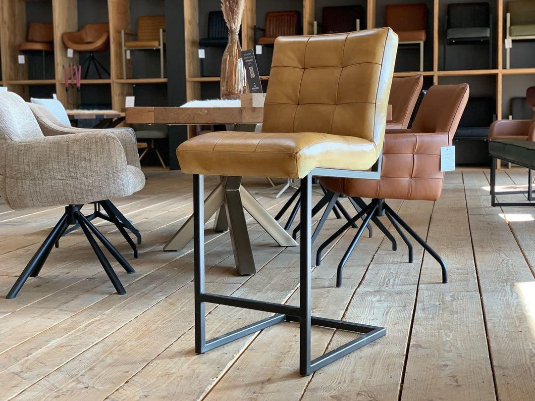 Thekenstuhl Leder Industrial in Mustard: Hochwertiger Thekenstuhl aus senfgelbem Leder mit gepolsterter Sitzfläche und gesteppter Rückenlehne, kombiniert mit einem stabilen schwarzen Metallgestell im Industrial-Stil, präsentiert auf einem Holzboden in einem modernen Möbelgeschäft mit weiteren Stühlen im Hintergrund.
